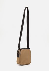 Brun canvas skuldertaske med en sort lynlås og rem. Firkantet form med glat tekstur og forstærkede hjørner. Har justerbar rem.