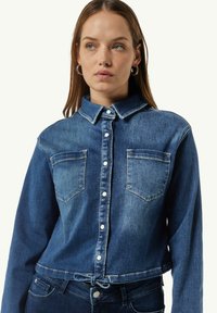 Denimhemd in Mittelblau mit zwei Brusttaschen, Knopfverschluss und cropped Design. Verfügt über einen Kragen und einen Kordelzug am Saum.