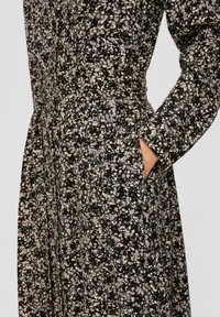 Zwarte bloemenjurk met lange mouwen, voorzien van een textuurpatroon in crème en beige. Inclusief zijzakken en een getailleerd ontwerp bij de taille.