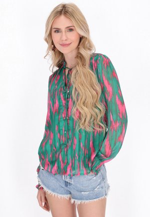 Blusa verde e rosa a motivi con maniche lunghe, chiusura a bottoni e polsini elastici, abbinata a pantaloni corti di jeans sfilacciati.