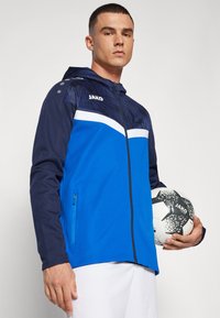 Blue and navy hooded jacket with a geometric pattern, white accents, and a zip pocket. Model holds a black and white soccer ball.