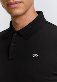 Pólo negro hecho de tejido texturizado, con una tapeta de tres botones y un logo distintivo bordado en blanco en el pecho.