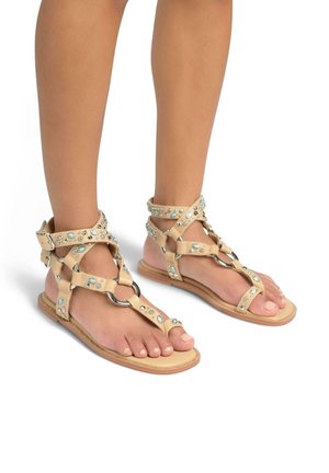 Beige leren sandalen met decoratieve blauwe edelstenen, meerdere banden en metalen cirkelaccenten; platte zool met lichte stikdetails.