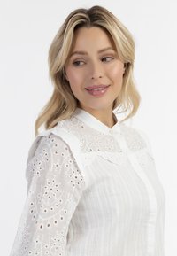 Witte blouse met geborduurde oogjesdetails, gestructureerde stof, een kraag en knoopsluiting, met lange mouwen en delicate accenten.