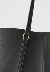 Sac fourre-tout en cuir noir avec une surface texturée, mettant en avant un accent en métal doré et un design simple et minimaliste.