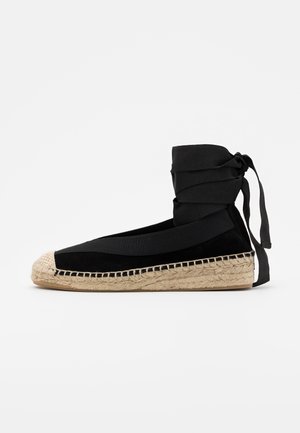 Espadrille plate noire avec semelle en jute tissée, orteil arrondi et ruban enroulé autour de la cheville noué à l'arrière sur un fond blanc.