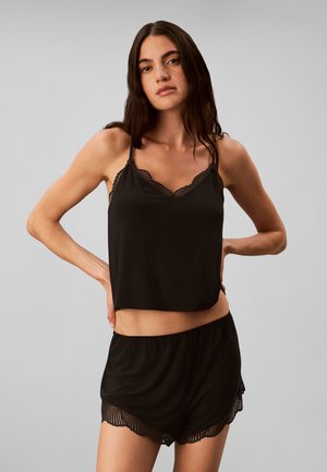 Mujer con cabello largo y oscuro que lleva una camiseta sin mangas negra con encaje y pantalones cortos a juego, de pie con las manos en las caderas contra un fondo liso.