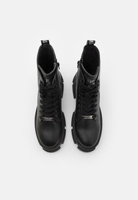 Paire de bottes de combat en cuir noir avec semelles épaisses, lacets, fermetures éclair latérales et étiquettes métalliques de marque sur fond blanc.