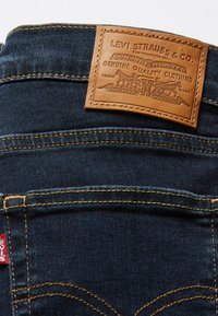Jeans in denim blu scuro con una patta in cuoio marrone con dettagli in rilievo, cuciture dorate e un'etichetta rossa sulla tasca posteriore.