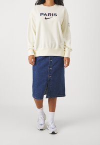 Persona vistiendo un sudadera color crema con "PARIS" y el logotipo de Nike, falda de mezclilla azul oscuro con botones en frente, calcetines blancos y zapatillas blancas.