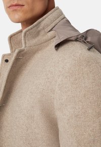 Hombre con abrigo de lana beige con cuello alto y capucha desmontable sujeta por un cordón en el hombro.