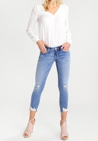 Blusa blanca de manga larga con escote en V, combinada con jeans ajustados de tiro bajo en un tono azul claro desgastados y sandalias de tacón en rosa pálido.