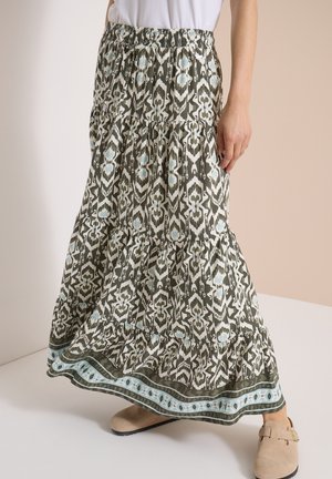 Kvinde iført et langt, mønstret skørt med grønne, hvide og blå geometriske designs, kombineret med beige slip-on sko.