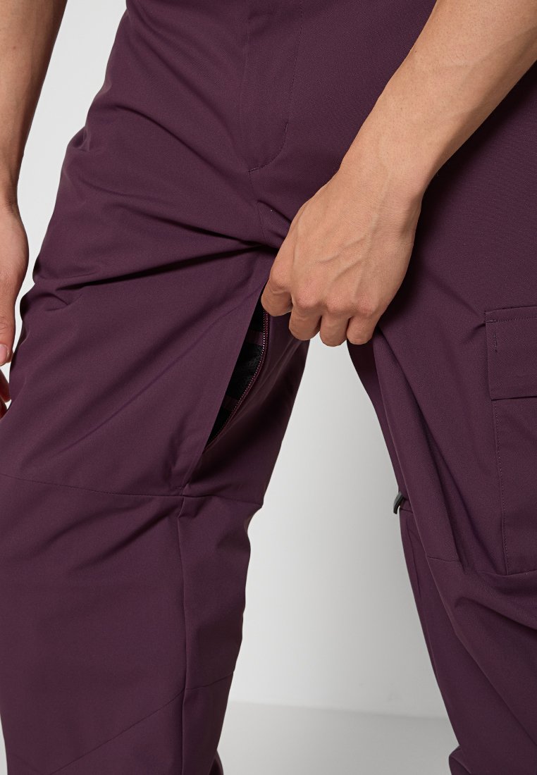 Primo piano di una mano che apre la cerniera laterale di pantaloni cargo di un colore viola scuro, mostrando un motivo a righe all'interno.