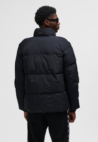 Sort pufferjakke med høj krave, quiltede sektioner og lange ærmer. Stoffet virker glat og mat uden synlige mønstre.