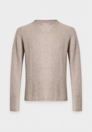 Homme adulte debout portant un pull beige, un jean bleu clair, une ceinture marron et des mocassins marron sur un fond clair uni.
