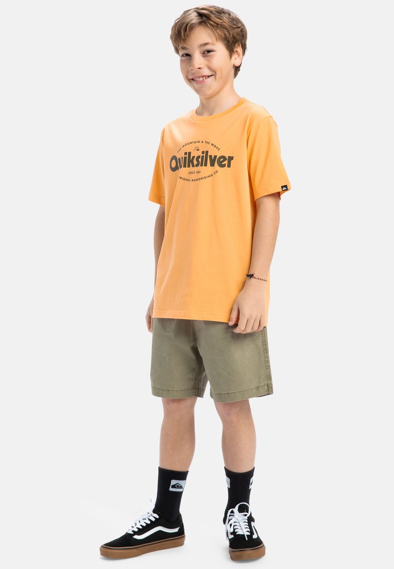 Oranssi puuvillapaita, jossa on "Quiksilver"-logo, yhdistettynä oliivinvihreisiin shortseihin, mustavalkoisiin urheilukenkiin ja mustiin sukkiin.