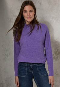 Pull en tricot côtelé violet avec un col montant et des manches longues, associé à un jean bleu foncé. La texture est douce et légèrement ajustée.