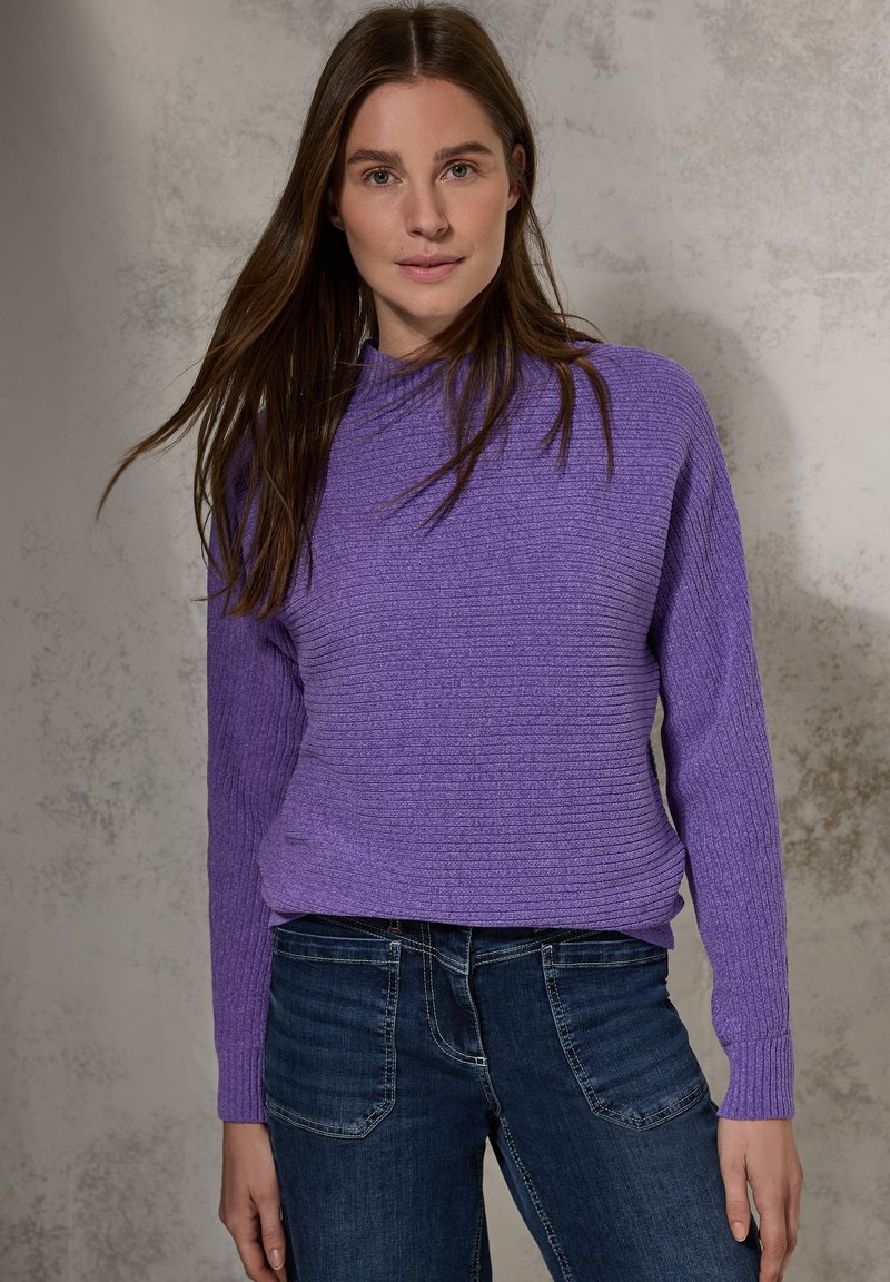 Pull en tricot côtelé violet avec un col montant et des manches longues, associé à un jean bleu foncé. La texture est douce et légèrement ajustée.