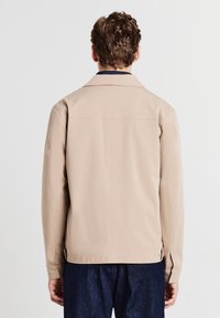 Giacca beige con colletto, maniche lunghe e una texture liscia. Presenta dettagli con spacco laterale e un design minimalista.