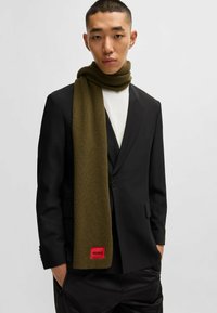 Sciarpa verde a coste con un'etichetta rossa, indossata sopra una camicia bianca e un giacca nera, con una texture liscia e un design a strati.