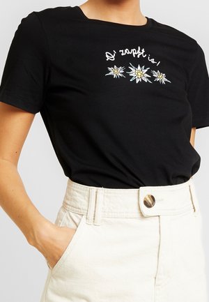 Persoon draagt een zwart T-shirt met witte en gele bloemgrafieken en tekst erboven, ingestopt in hoog uitgesneden lichtbeige broek met knoop.