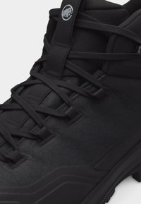 Bocanc de drumeție negru înalt, cu țesătură texturată detaliată, talpă groasă și logo rotund pe limbă, lângă șireturi negre strâns legate.