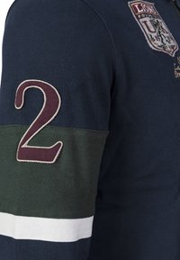Granatowa koszula z długim rękawem z zielonymi i białymi pasami na rękawie, dużą bordową cyfrą "2" oraz wyszywanym herbem z tekstem na piersi.