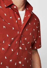 Chemise à manches courtes couleur rouille avec petits motifs de silhouettes de chiens blancs imprimés partout, portée par-dessus un t-shirt blanc à col rond.