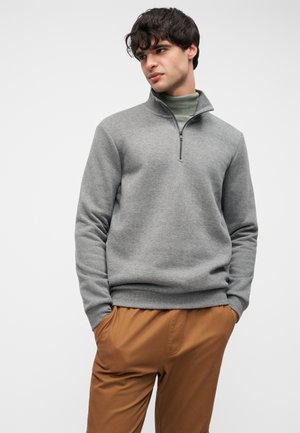 Maglione grigio con chiusura a zip, collo alto, polsini e orlo a coste; abbinato a pantaloni marroni. Tessuto morbido, vestibilità casual, design minimale.