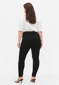 Jeans skinny de tiro alto en negro con una textura suave, dos bolsillos traseros y un diseño ajustado, combinados con una blusa blanca y tacones negros.