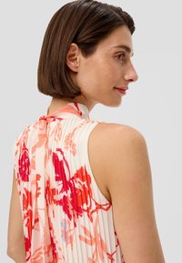 Plissiertes ärmelloses Oberteil aus weichem Stoff, mit einem Blumenmuster in Rot- und Rosatönen auf einem hellen Hintergrund. Mit Krawatten am Nackenverlauf hinten.