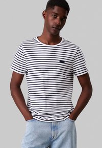 Cameriere giovane, indossando una maglietta a righe bianche e blu scuro e jeans azzurri, in piedi con le mani nelle tasche contro uno sfondo neutro.