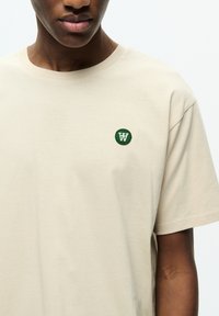 Beige bomulds t-shirt med korte ærmer, der har et grønt broderet cirkulært logo på venstre brystområde. Glat tekstur, rund halsudskæring.