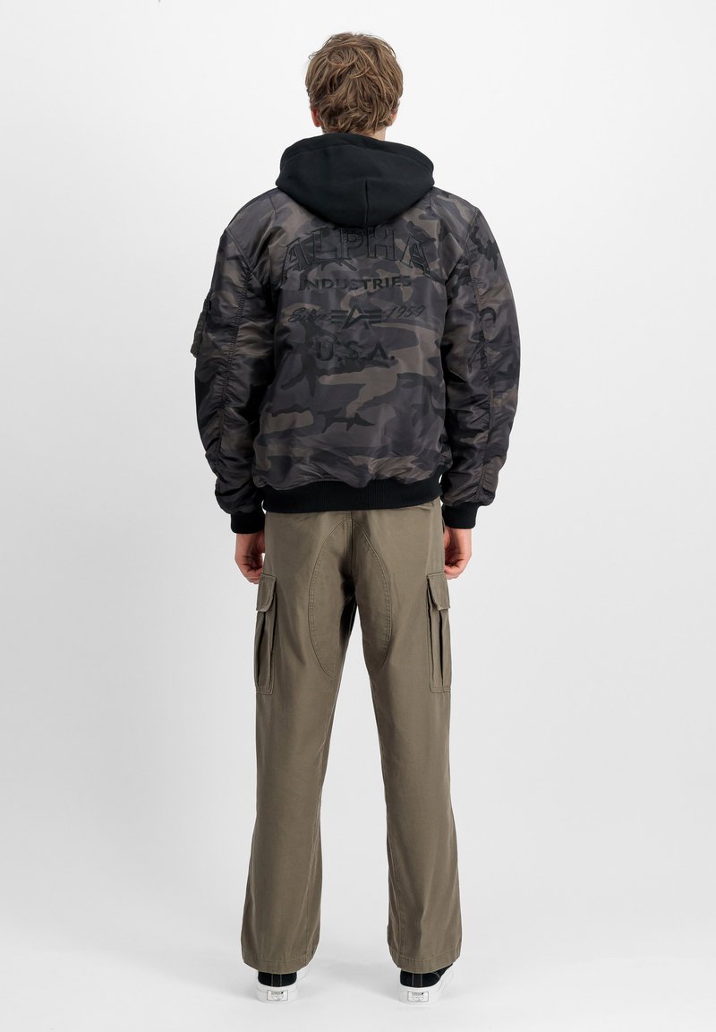 Giacca bomber mimetica nera e grigia con polsini a costine, felpa con cappuccio sotto e pantaloni cargo verde oliva con tasche laterali.