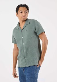 Camicia verde a maniche corte con bottoni, in tessuto traforato, con una tasca sul petto e un marchio discreto sopra. Indossata con jeans blu.