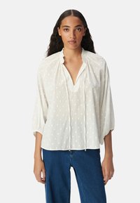 Blusa bianca con pois neri, scollatura a V e colletto arricciato. Presenta maniche lunghe a sbuffo e un tessuto leggero e testurizzato.