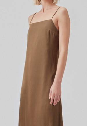 Kobieta ubrana w brązową sukienkę na ramiączkach, z cienkimi, spaghetti paskami, stojąca na tle jednolitego jasnoszarego tła.