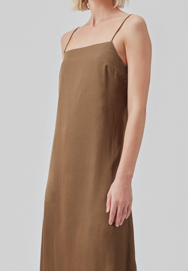 Femme portant une robe nuisette marron sans manches avec de fines bretelles spaghetti, debout devant un fond uni gris clair.