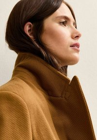 Jonge vrouw met bruin haar die een hooggesloten, gestructureerde camelkleurige jas draagt, terwijl ze bedachtzaam naar rechts kijkt tegen een eenvoudige achtergrond.