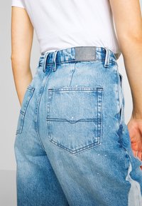 Jeans in denim blu chiaro con zone sbiadite e dettagli di schizzi di vernice bianca. Presentano un'etichetta in vita e due tasche posteriori.