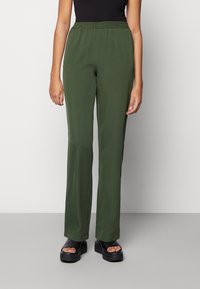 Groene, ontspannen-fit broek met een elastische tailleband en gladde textuur. Het ontwerp heeft rechte pijpen en een lichte glans, gecombineerd met zwart schoeisel.