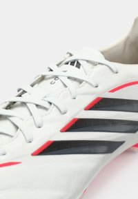 Λευκό αθλητικό παπούτσι με γκρίζες και μαύρες λωρίδες, έντονες κόκκινες λεπτομέρειες, υφή στη πάνω πλευρά και παραδοσιακό σχέδιο με κορδόνια.