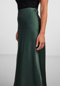 Falda de satén verde con una textura suave y un ligero brillo. Presenta una cintura ajustada y una silueta fluida. Mano apoyada sobre la tela.