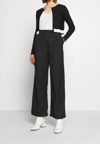 Cárdigan corto negro con tejido texturizado, combinado con pantalones de pierna ancha negros de cintura alta. Top blanco ajustado debajo y botines blancos.