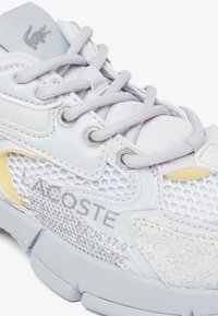 Sneaker grigio chiaro con una combinazione di texture in mesh e suede, con lacci e un logo sul lato. Accenti gialli e bianchi visibili.