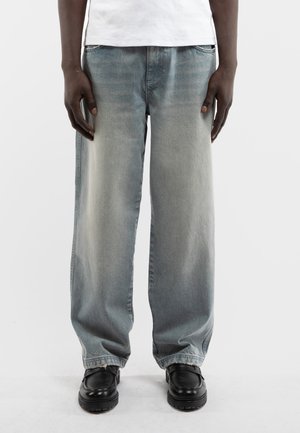 Osoba ubrana w sprane niebieskie szerokie dżinsy, czarne buty i białą koszulę, stojąca na tle jednolitego białego tła.