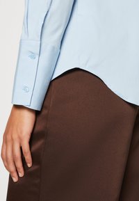 Main reposant sur le côté portant une chemise bleu clair à manches longues avec un poignet boutonné et un pantalon marron avec un ourlet de chemise courbé par-dessus.