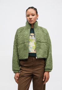 Zelená fleecová bunda s texturovaným, huňatým povrchem, vysokým límcem a zapínáním na zip; nošená přes tričko s potiskem a hnědé kalhoty.