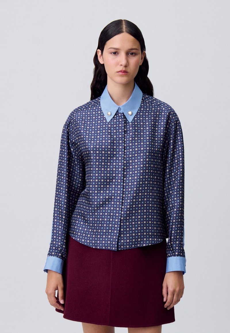 Camisa azul marino con un cuello geométrico azul claro, que presenta un patrón de cuadrícula con pequeños cuadrados y dos botones similares a perlas en el cuello.
